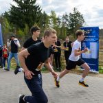 DSC_5814 Na zdjęciu widać trzech biegnących młodych ludzi w czarno-białych strojach. W tle znajduje się las oraz niebieski baner reklamowy. Po lewej stronie widać nadmuchiwany łuk startowy w biało-różowych barwach.