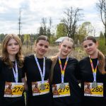 DSC_5819 Cztery osoby w czarnych koszulkach z numerami startowymi i medalami. W tle drzewa i pochmurne niebo. Uczestnicy stoją blisko siebie.