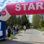 DSC_5824 Biegacz przekracza linię startu pod różowo-białym łukiem z napisem START. W tle zielone drzewa i niebieskie niebo. Kibice stoją po bokach drogi.