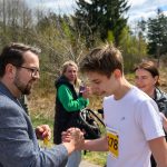 DSC_5830 Na zdjęciu widać ludzi na świeżym powietrzu klaskających sobie w dłonie. Młodzieniec w białej koszulce ma żółty numer startowy. W tle są drzewa i zarośla, a niebo jest częściowo zachmurzone.