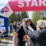 DSC_5838 Na zdjęciu osoba wręcza medal biegaczowi pod różowym łukiem z napisem START. W tle grupa ludzi i drzewa. Słońce świeci, tworząc jasną atmosferę.