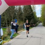 DSC_5840 Biegacz w czarnym stroju kończy bieg pod różowo-niebieskim balonem. Droga otoczona zielonym lasem. Na boku stoi fotograf.