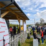 DSC_5856 Na zdjęciu grupka ludzi na zewnątrz przy drewnianym zadaszeniu. Tło to błękitne niebo z chmurami i drzewa. Widoczne są banery z logo Mazowsze.