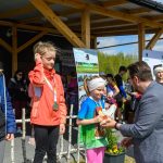 DSC_5867 Na zdjęciu są trzy dzieci z medalami, ubrane kolorowo, odbierające nagrody. W tle widoczna jest drewniana konstrukcja i baner. Tło jest zielone z niebieskim niebem, a kompozycja jest żywa i radosna.