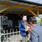 DSC_5869 Na zdjęciu jest osoba w granatowej kurtce, która otrzymuje trofeum na zewnątrz. W tle są drewniana wiata i baner z napisem „Mazowsze”. Obok stoją osoby, a pogoda jest słoneczna.