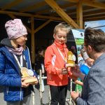 DSC_5871 Dwie dziewczynki otrzymują trofea na podium, ubrane w kolorowe kurtki. W tle drewniana altana i ludzie. Słoneczny dzień.