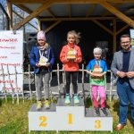 DSC_5875 Trzy dziewczynki na podium z trofeami na tle drewnianego budynku. Niebieskie niebo, plakaty i zielona trawa w tle. Obok stoi mężczyzna.