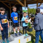 DSC_5876 Trzech chłopców stoi na podium w niebieskich strojach sportowych. Otrzymują medale i trofea. W tle jest drewniana pergola i zieleń.
