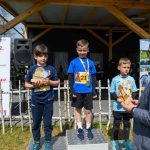 DSC_5879 Trzech chłopców na podium z pucharami w rękach. Tło z logiem Mazowsze i drewnianym zadaszeniem. Mężczyzna w marynarce wręcza nagrody.