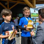 DSC_5881 Dwoje dzieci w niebieskich koszulkach trzyma trofea. W tle jasne drewniane zadaszenie i reklama z zielonym krajobrazem. Mężczyzna w okularach rozmawia z jednym z chłopców.