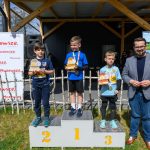 DSC_5885 Chłopcy stoją na podium z trofeami. Obok nich mężczyzna w marynarce. Tło to drewniana konstrukcja i plakaty reklamowe.
