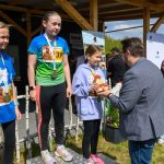 DSC_5888 Na zdjęciu widnieją trzy młode dziewczyny na podium w sportowych strojach. Każda ma medal, a jedna z nich trzyma trofeum. W tle widać drewnianą konstrukcję i niebo z chmurami.