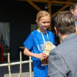 DSC_5891 Na zdjęciu dziecko w niebieskiej koszulce otrzymuje trofeum. W tle drewniana konstrukcja i głośnik. Dzień jest słoneczny.