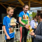 DSC_5892 Dwie młode dziewczyny na podium z pucharami, ubrani w kolorowe, sportowe koszulki. Obok nich stoi mężczyzna w marynarce, który wręcza nagrody. Tło z drewnianą konstrukcją i plakatami.