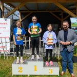 DSC_5895 Trzy dziewczynki stoją na podium z trofeami, obok mężczyzna. Tło to drewniana konstrukcja i banery. Kolory jasne, wiosenne.