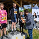DSC_5900 Trzech chłopców stoi na podium. Jeden mężczyzna wręcza im nagrody. W tle drewniana konstrukcja i baner.
