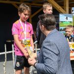 DSC_5902 Na zdjęciu kilku chłopców stoi na podium. Jeden z nich, w różowej koszulce, otrzymuje nagrodę od mężczyzny. W tle widoczne zielone drzewa i zadaszona konstrukcja.