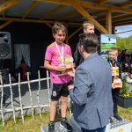 DSC_5903 Na zdjęciu jest ceremonia wręczenia nagród z uśmiechniętymi dziećmi na podium. Dziecko w różowej koszulce trzyma trofeum. Tło to drewniana konstrukcja i kwiaty w doniczce.
