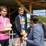 DSC_5904 Na zdjęciu widać dwóch chłopców z medalami i nagrodami na tle drewnianej wiaty. Mężczyzna im gratuluje, a za nimi widać uczestników biegu. Tło zawiera niebo, drzewa i osoby w sportowych strojach.