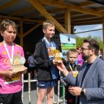 DSC_5905 Chłopcy na podium z trofeami w otoczeniu dorosłego. Różne barwne ubrania, tło z plakatami. Słoneczny dzień na zewnątrz.