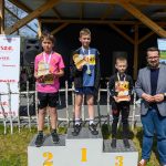DSC_5908 Trzech chłopców z medalami stoi na podium; liczby 1-3 są żółte. Po lewej białe tło z czerwonym napisem Mazowsze. Tło z drewnianą altaną i zieloną trawą.