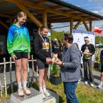 DSC_5910 Na podium stoją dwie młode dziewczyny w sportowych strojach, jedna w zielono-niebieskiej bluzie. Mężczyzna w marynarce wręcza jej nagrodę. W tle widoczna jest polska flaga i grupa osób.