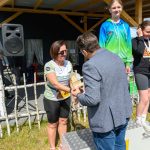 DSC_5912 Na zdjęciu widać dekorację zwycięzców na podium. Kobieta odbiera nagrodę od mężczyzny na tle baneru z napisem Mazowsze. Tło jest ciemne z elementami drewna i zielenią trawy.