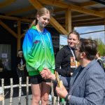 DSC_5914 Na zdjęciu widać młodą dziewczynę na podium w zielono-niebieskiej bluzie. Otrzymuje nagrodę od mężczyzny. W tle drewniana altana i baner.
