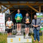 DSC_5918 Na zdjęciu widać ceremonii wręczenia nagród na podium. Trzy osoby trzymają trofea, stojąc na tle drewnianego budynku. Jasne kolory ubrań kontrastują z ciemnym tłem.