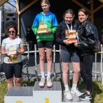DSC_5922 Trzy osoby stoją na podium, trzymają trofea. Tło to drewniana konstrukcja i trawa. Dominuje kolor szary i zielony.