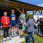 DSC_5924 Trzech chłopców na podium w sportowych ubraniach z medalami na szyi. Tło drewniane z zadaszeniem, niebo zachmurzone. Obok wręczanie nagród.
