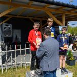 DSC_5925 Trzech chłopców na podium otrzymuje medale i trofea. Mężczyzna wręcza trofeum. Tło stanowi drewniany budynek i transparenty.