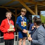 DSC_5927 Na zdjęciu dwaj chłopcy otrzymują nagrody sportowe na podium. Jeden ma czerwoną bluzę, drugi niebieski strój sportowy. W tle drewniana konstrukcja i zieleń.