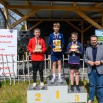DSC_5929 Trzech chłopców stoi na podium z trofeami. Tło to drewniana altana z plakatami i zieloną trawą. Słoneczny dzień z niebieskim niebem.
