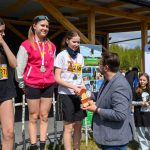 DSC_5933 Na zdjęciu trzy kobiety na podium otrzymują medale. Tło stanowi drewniana konstrukcja i baner z logo Gminy Dobre. W tle widoczne są drzewa i kilka osób.