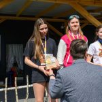 DSC_5936 Trzy młode osoby stoją na podium, trzymając trofea. Tło to drewniana konstrukcja ze sceną i głośnikiem. Jest słoneczny dzień.