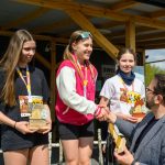 DSC_5937 Trzy młode dziewczyny otrzymują nagrody na podium sportowym. Mają na sobie medale, jedna jest w różowej bluzie. W tle drewniane zadaszenie i zieleń.