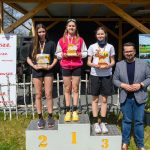 DSC_5942 Trzy młode kobiety stoją na podium, trzymają trofea. Mężczyzna po prawej w marynarce. Tło to drewniana altana i bannery.