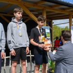 DSC_5945 Trzech młodych sportowców z medalami stoi na podium, w tle drewniana konstrukcja. Jeden z nich odbiera nagrodę od mężczyzny. Słoneczny dzień, kilka osób obserwuje w tle.