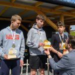 DSC_5948 Trzech chłopców w bluzach i medalach trzyma trofea. Otrzymują je od mężczyzny w marynarce. W tle drewniana altana i zieleń.