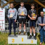 DSC_5950 Trzech młodych sportowców stoi na podium, trzymając trofea. Obok nich stoi mężczyzna w marynarce. Tło to drewniana konstrukcja i banery.