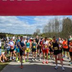 DSC_5968 Grupa biegaczy stoi na linii startu pod różowym łukiem. W tle są drzewa i niebieskie niebo. Po lewej widać biało-niebieski namiot.