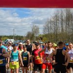 DSC_5970 Grupa biegaczy stoi na linii startu biegu na świeżym powietrzu. Ubrani w kolorowe stroje sportowe, otoczeni zielenią lasu. Słoneczny dzień, na niebie widoczne są chmury.