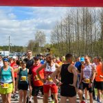 DSC_5971 Na zdjęciu grupa biegaczy w kolorowych strojach stoi na linii startu na zewnątrz. Tło stanowią drzewa i lekko pochmurne niebo. Widoczne są numery startowe i kibice w oddali.