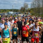 DSC_5972 Biegacze w kolorowych strojach czekają na start na tle lasu i nieba. Uliczka pełna ludzi. Słoneczny dzień, dużo zieleni.
