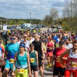 DSC_5974 Grupa osób w kolorowych ubraniach czeka na start biegu. Za nimi widać drzewa i kilka zaparkowanych aut. Nad nimi rozciąga się niebieskie niebo z chmurami.