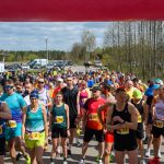 DSC_5976 Grupa biegaczy na starcie maratonu, kolorowe stroje i numery startowe. Tło to las i parking z samochodami. Pogoda jest słoneczna.