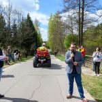 DSC_5989 Na zdjęciu grupa ludzi na drodze w parku, mężczyzna mówi do mikrofonu. Obok czerwony quad, w tle drzewa i niebo. Dzień jest słoneczny.