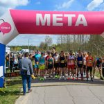 DSC_5990 Biegacze stoją pod różowo-białym łukiem z napisem META na tle nieba i drzew. Wydarzenie sportowe odbywa się na asfalcie. Po lewej stronie widoczny zegar czasu.