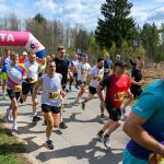DSC_6015 Na zdjęciu grupa biegaczy ubranych w kolorowe koszulki biegnie po asfaltowej drodze. W tle znajduje się las i różowy łuk z napisem META. Jest słoneczny dzień z lekkim zachmurzeniem.